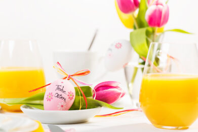 Rosa Ostereier mit der Aufschrift Frohe Ostern, Tulpen und Gläser mit Orangensaft auf einem hellen Frühstückstisch.